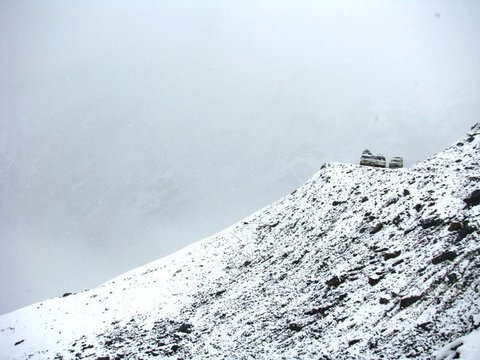 Two small vehicles perch on a snow-dusted rocky ridge that drops away into a wall of grey cloud.