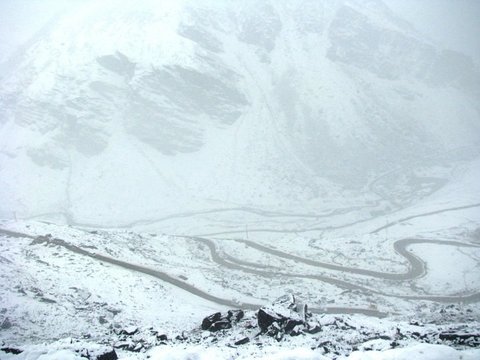 A mountain road loops in long switchbacks across a snow-covered valley floor, the slopes above dissolving into mist.