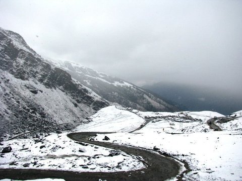 A wet black road curves between snow-covered slopes toward a cluster of huts, dark ridges and a cloud-filled valley beyond.