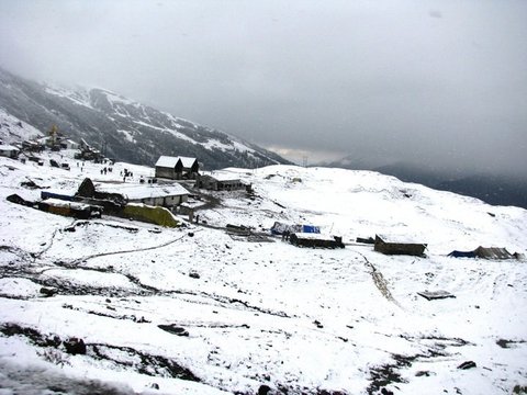 Snow blankets a scatter of stone huts, tents and tin roofs on a high slope, a snow-streaked ridge behind under a heavy grey sky.