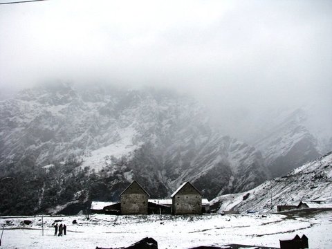 Two gabled stone buildings sit under fresh snow at the foot of a cloud-wrapped mountainside, a few small figures on the slope nearby.