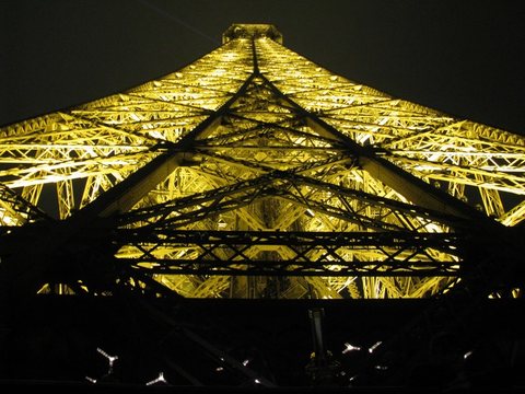The iron lattice of the Eiffel Tower seen from directly below at night, lit gold against a black sky.