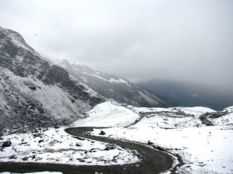 A dark mountain road curving through fresh snow towards a valley filled with storm cloud.