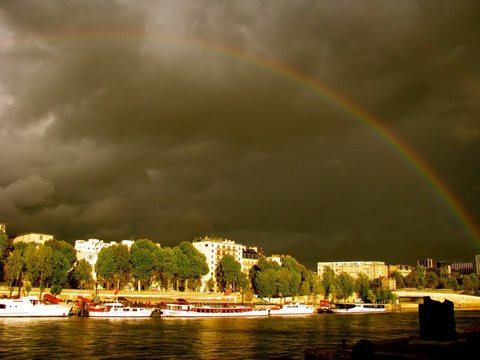 A rainbow arcing through dark storm clouds above sunlit trees and moored boats on the Seine.