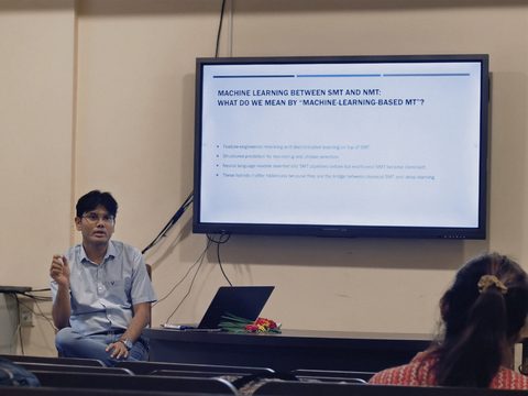 Dr. Pinaki Bhaskar speaking, seated beside a screen showing the slide “Machine learning between SMT and NMT”.